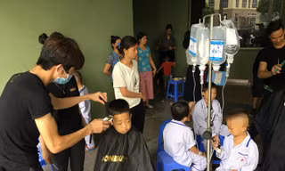 Nhiều bệnh nhi còn lủng lẳng bình dịch truyền xếp hàng ngồi chờ được cắt tóc. Ảnh: Đức Long