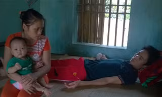 Chị Hiên bế đứa con út ngồi cạnh chồng bị tai nạn dẫn đến mất khả năng lao động