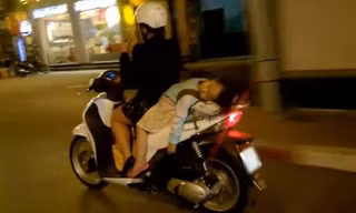 Em bé không được đội mũ bảo hiểm, nằm ngủ gật trên xe của mẹ.