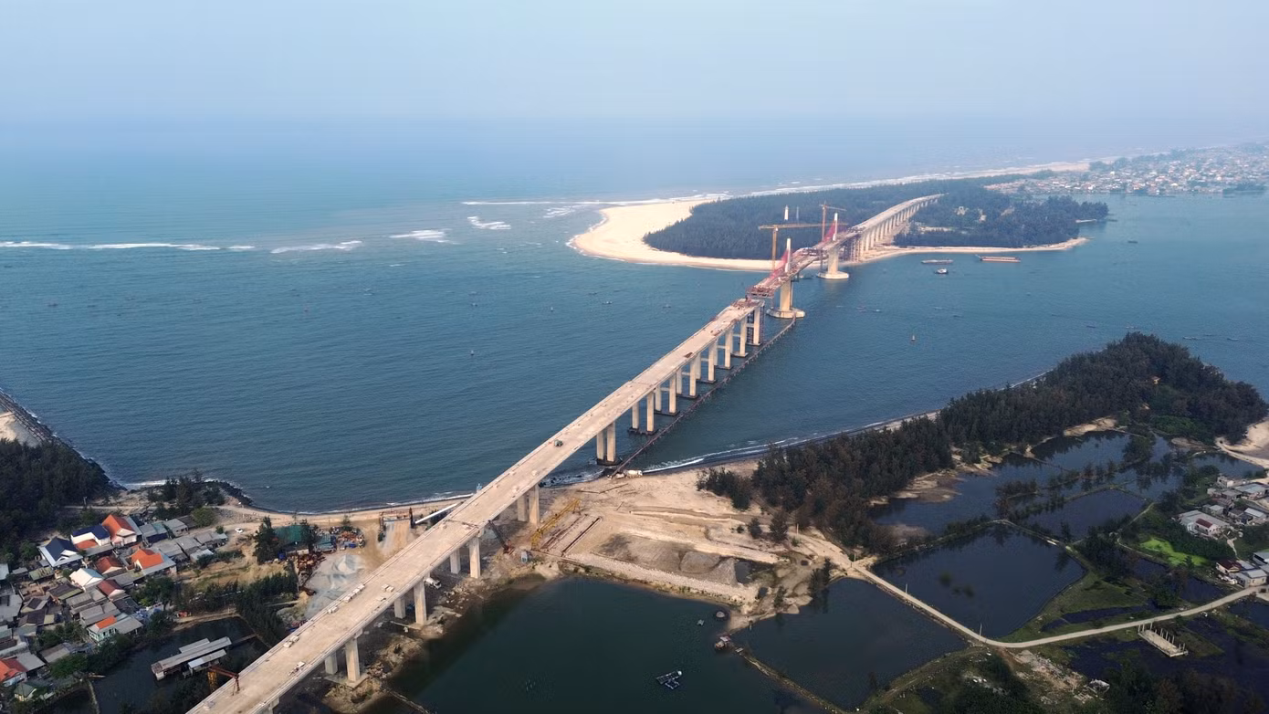 Nhìn từ trên cao, cây cầu như một dải lụa đỏ mềm mại vắt qua cửa biển Thuận An, nổi bật giữa nền trời và mặt biển xanh.
