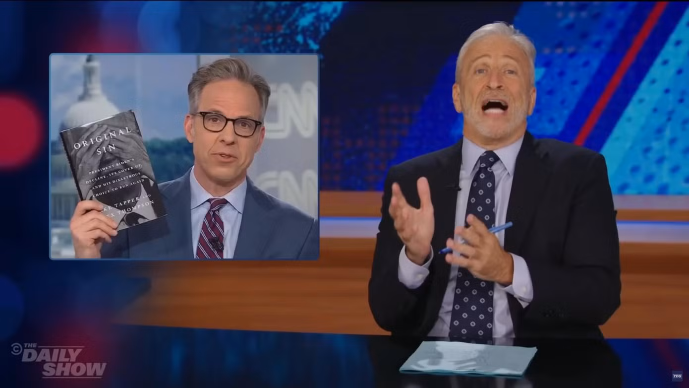 Jon Stewart chế giễu Jake Tapper vì liên tục giơ cuốn sách mới lên trước ống kính trong lúc dẫn chương trình trên CNN. Ảnh: The Daily Show.