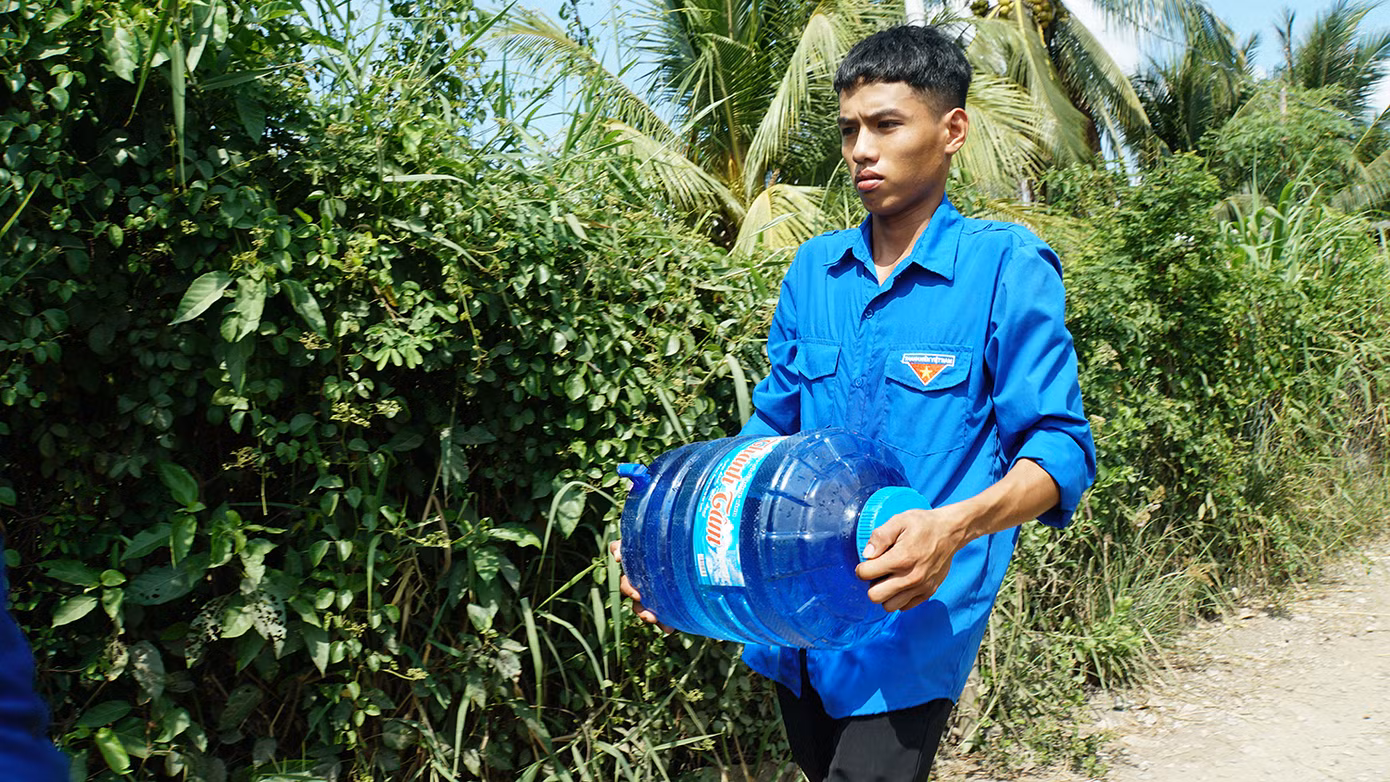 Nhà bà Trinh cách UBND xã hơn cây số, tuy nhiên để đến nơi phải đi trong con hẻm chưa đầy 1m, ngang vừa người, xe gắn máy phải chạy chậm, sâu bên trong. Trong ảnh, đoàn viên thanh niên mang nước đến tận nhà bà Trinh.